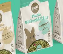 Drei Verpackungen von MUCKI Kleinnagerfutter mit den Sorten 'Menü Blütenblätter' und 'Menü Multi Mix' auf grünem Hintergrund