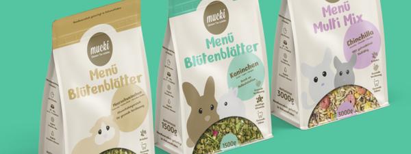 Drei Verpackungen von MUCKI Kleinnagerfutter mit den Sorten 'Menü Blütenblätter' und 'Menü Multi Mix' auf grünem Hintergrund